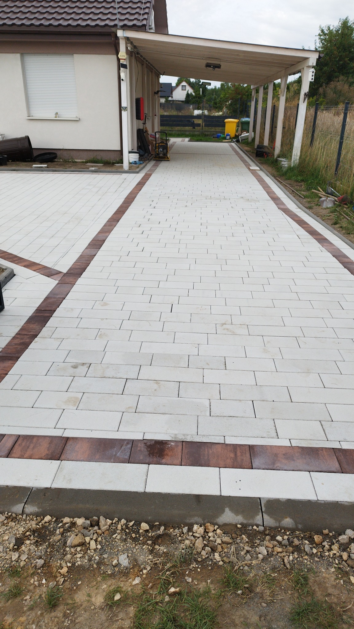 Ułożona kostka brukowa w odcieniach szarości i brązu tworząca wzór na podjeździe. Widoczne obrzeża i fragment domu z zadaszeniem. Prace brukarskie w toku.