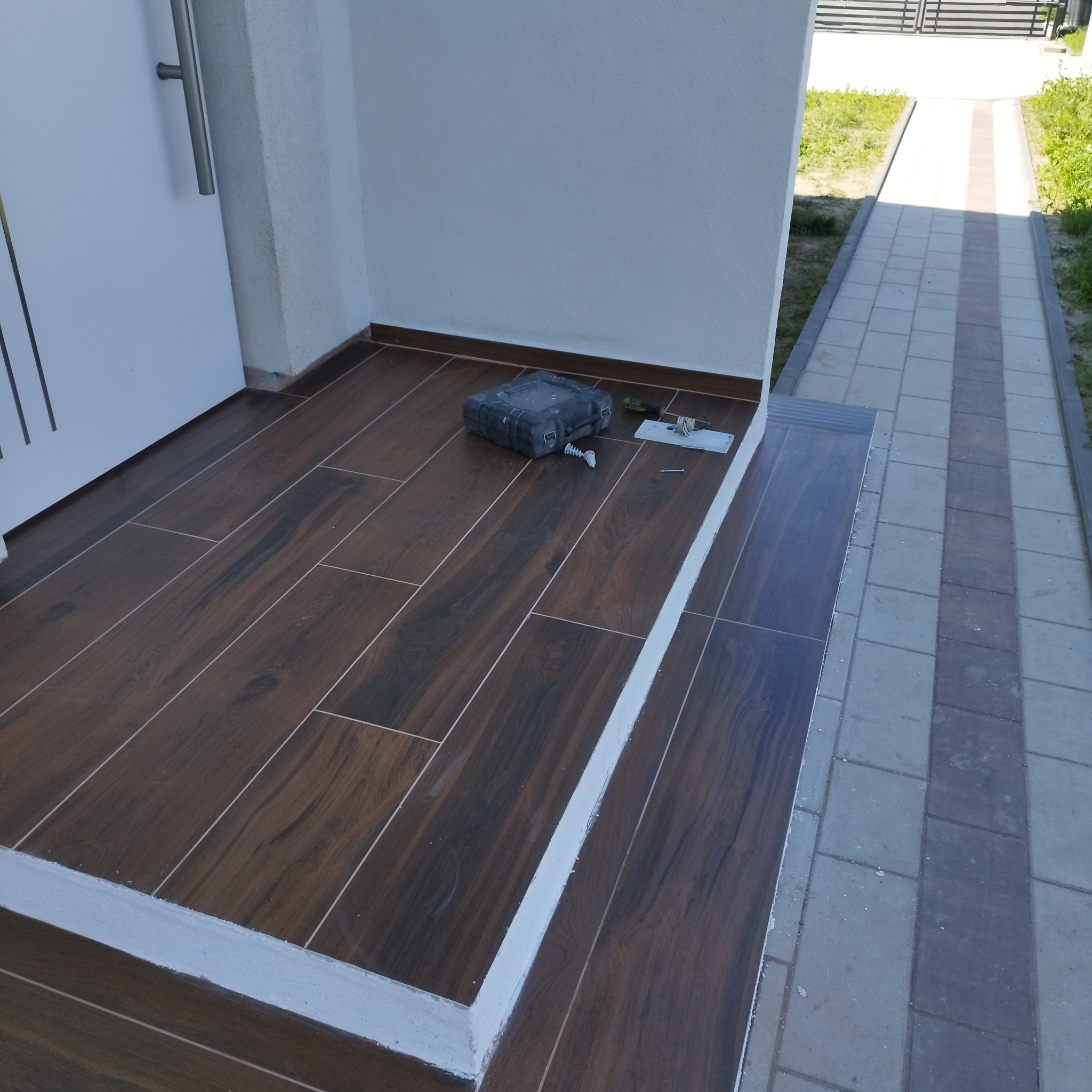 Wykończony taras z brązowych płytek imitujących drewno, z widoczną skrzynką narzędziową i akcesoriami montażowymi. Białe obramowanie i kostka brukowa na zewnątrz.