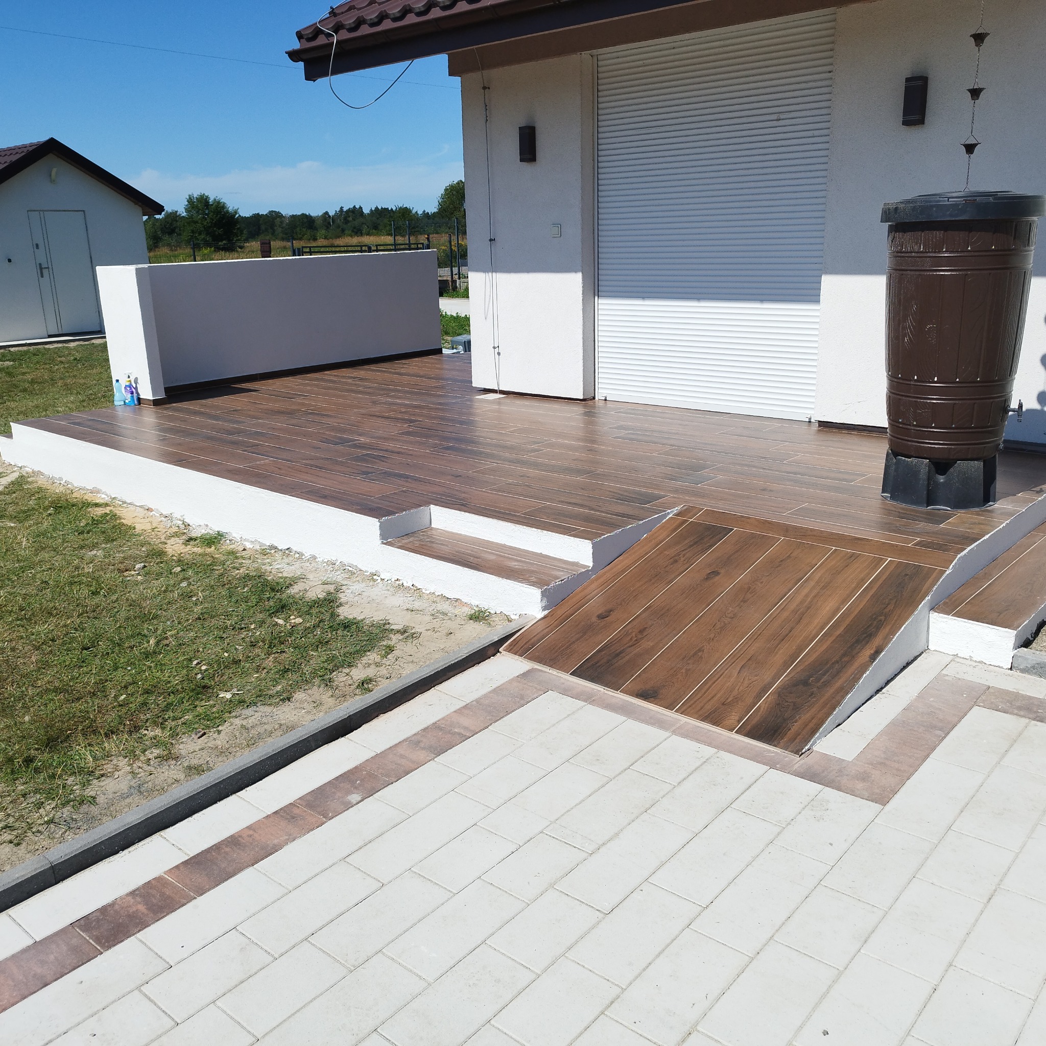 Wykończony taras z brązowych płytek imitujących drewno, z rampą i schodami. Obok biała kostka brukowa z brązowymi akcentami. W tle budynek z roletami.
