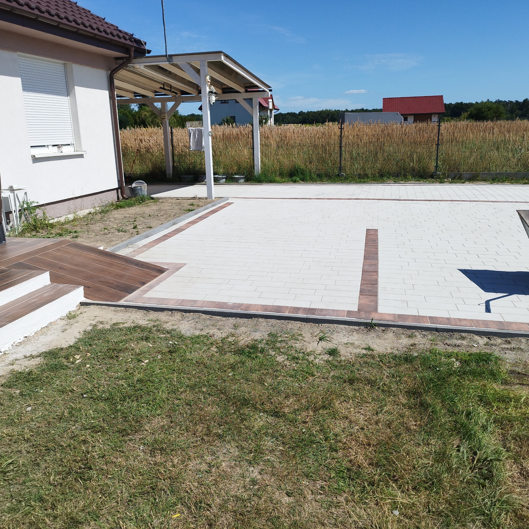 Jasna kostka brukowa z brązową obwódką na podjeździe domu jednorodzinnego w słoneczny dzień. Widoczny fragment tarasu i zieleń w tle.