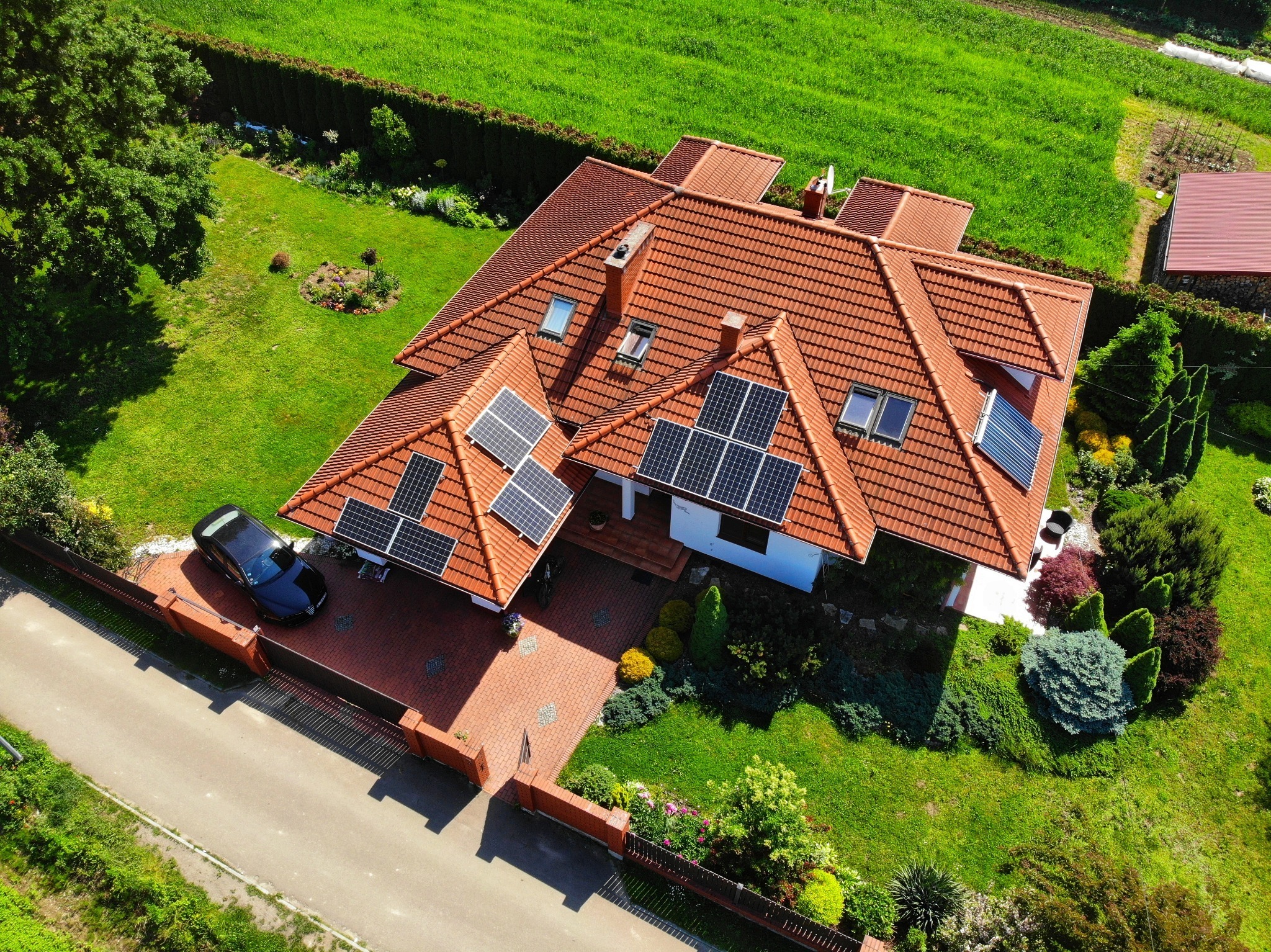 Widok z lotu ptaka na dom z czerwoną dachówką i panelami słonecznymi na dachu, zadbany ogród z trawą i drzewami, czarny samochód na podjeździe z kostki brukowej.