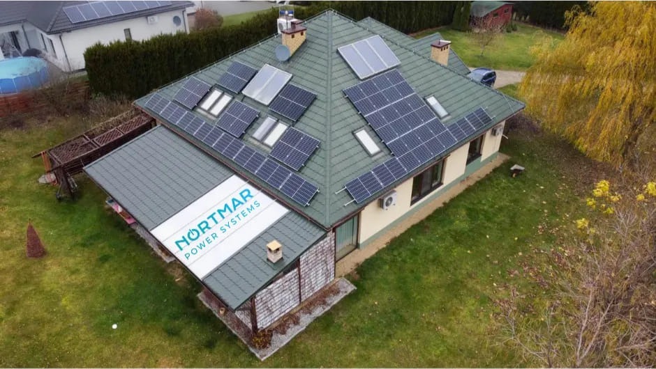 Dom jednorodzinny z zielonym dachem pokrytym panelami słonecznymi różnej wielkości i kształtu, widoczne okna dachowe, klimatyzator na ścianie, a na dobudówce logo firmy Nortmar Power Systems.