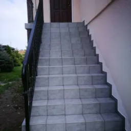 Zewnętrzne schody wejściowe wyłożone szarymi płytkami, z czarną, metalową balustradą i drzwiami wejściowymi w tle.