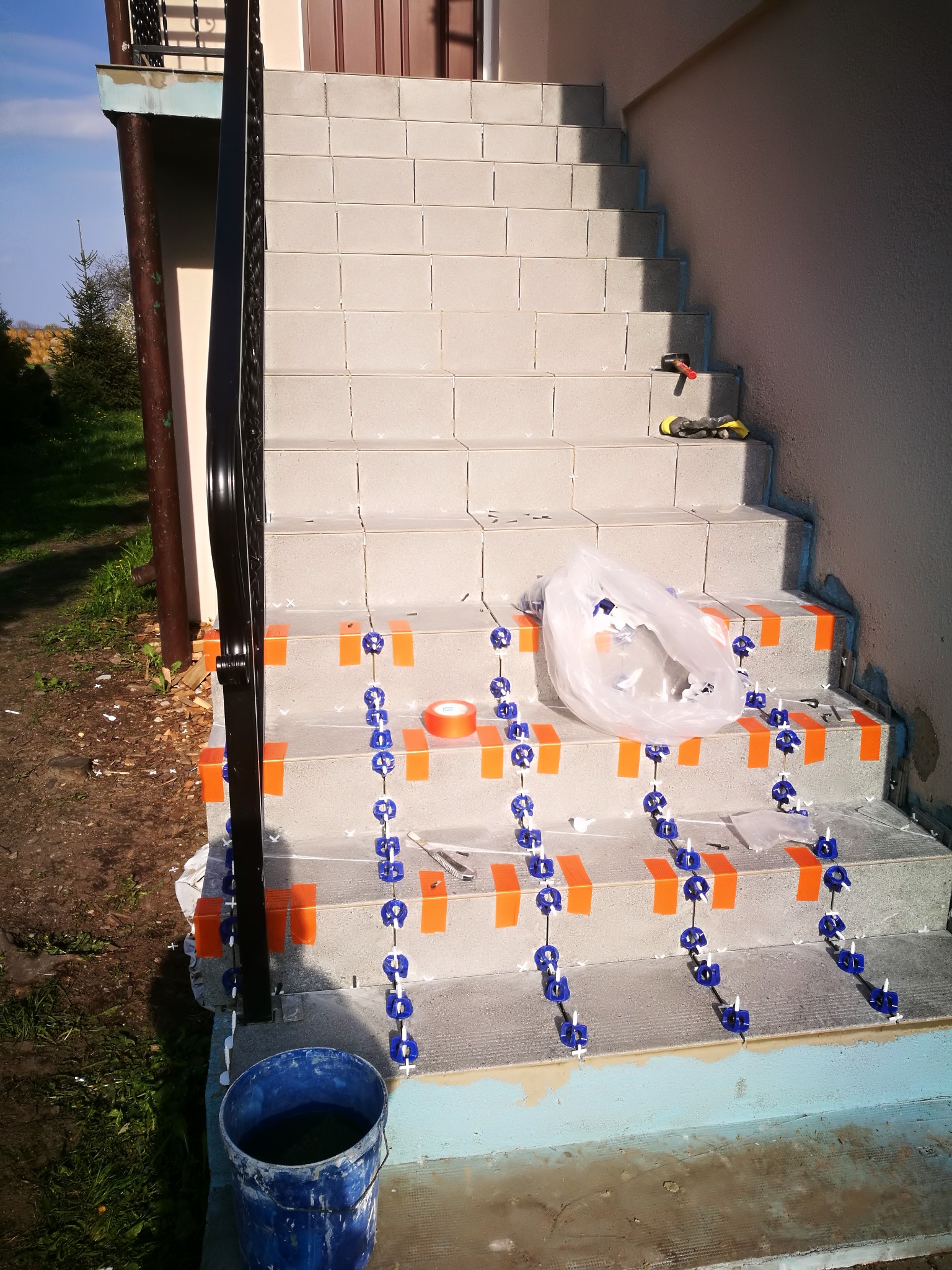 Schody zewnętrzne w trakcie układania płytek, z użyciem pomarańczowej taśmy i niebieskich klipsów poziomujących, widoczne narzędzia i wiadro z wodą.