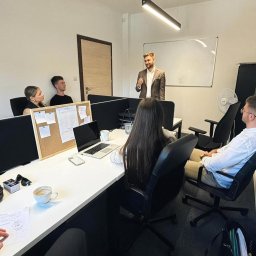 Na Miarę Nieruchomości - Mężczyzna w brązowej marynarce prezentuje coś grupie osób w biurze z białymi biurkami, laptopem, korkową tablicą i kubkiem kawy.