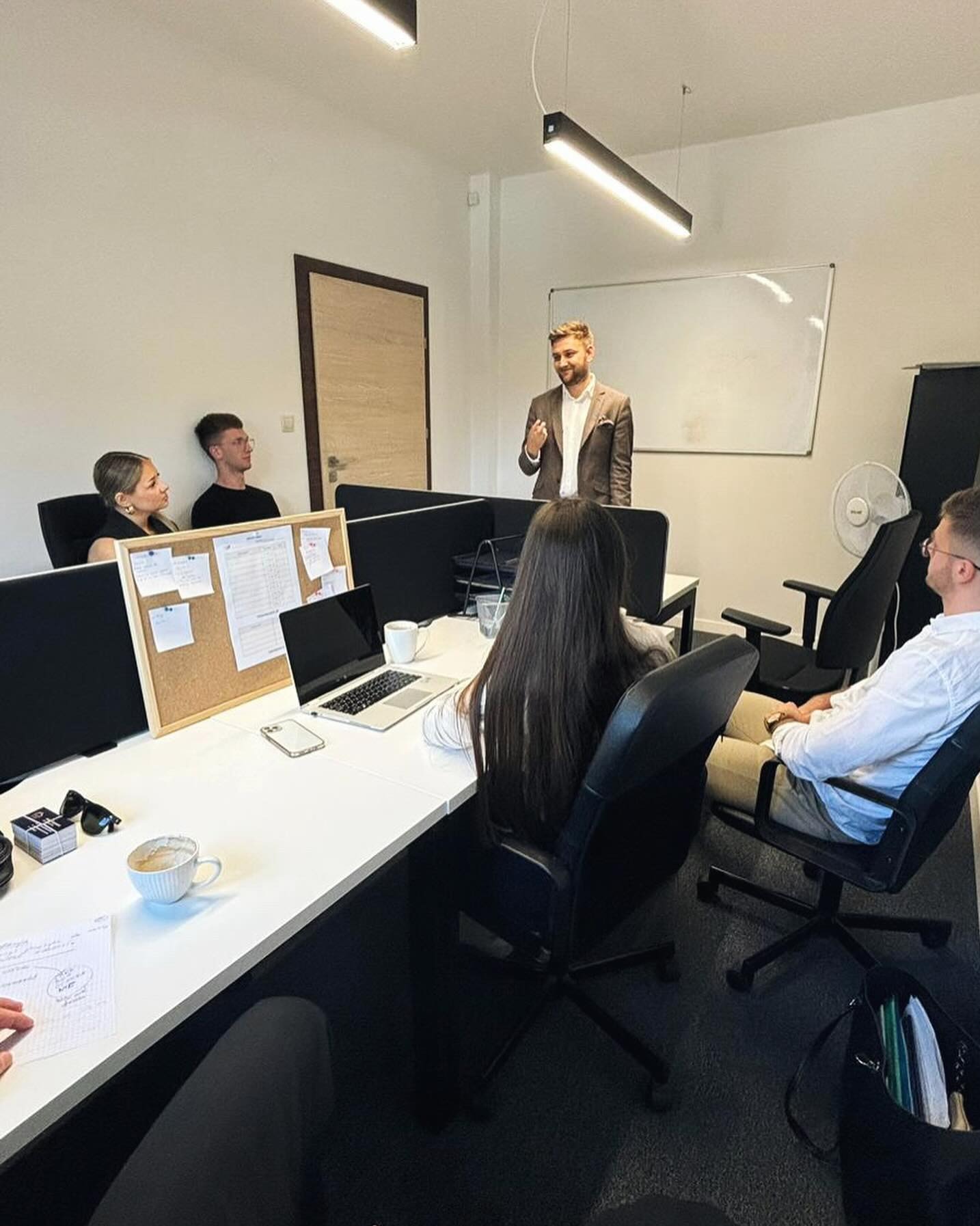 Mężczyzna w brązowej marynarce prezentuje coś grupie osób w biurze z białymi biurkami, laptopem, korkową tablicą i kubkiem kawy.