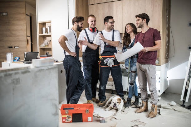 Grupa remontowa w roboczych kombinezonach omawia projekt z klientką w remontowanym pomieszczeniu kuchennym, na podłodze leży buldog angielski, a obok stoi pomarańczowa skrzynka z narzędziami.