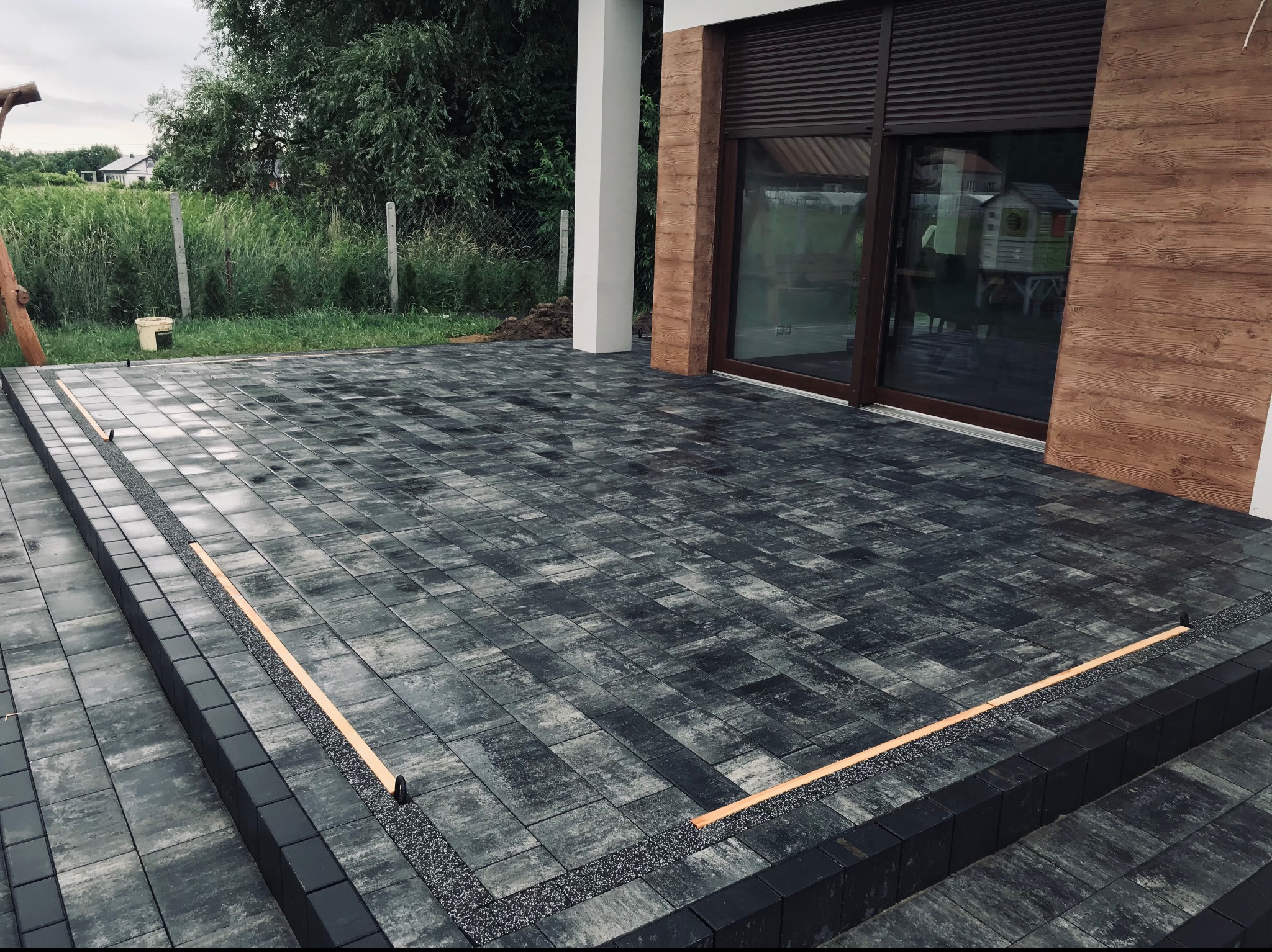 Nowoczesny taras z ciemnoszarej kostki brukowej, wykończony drobnym żwirem i obrzeżami, przylegający do domu z drewnianą elewacją i dużymi oknami.
