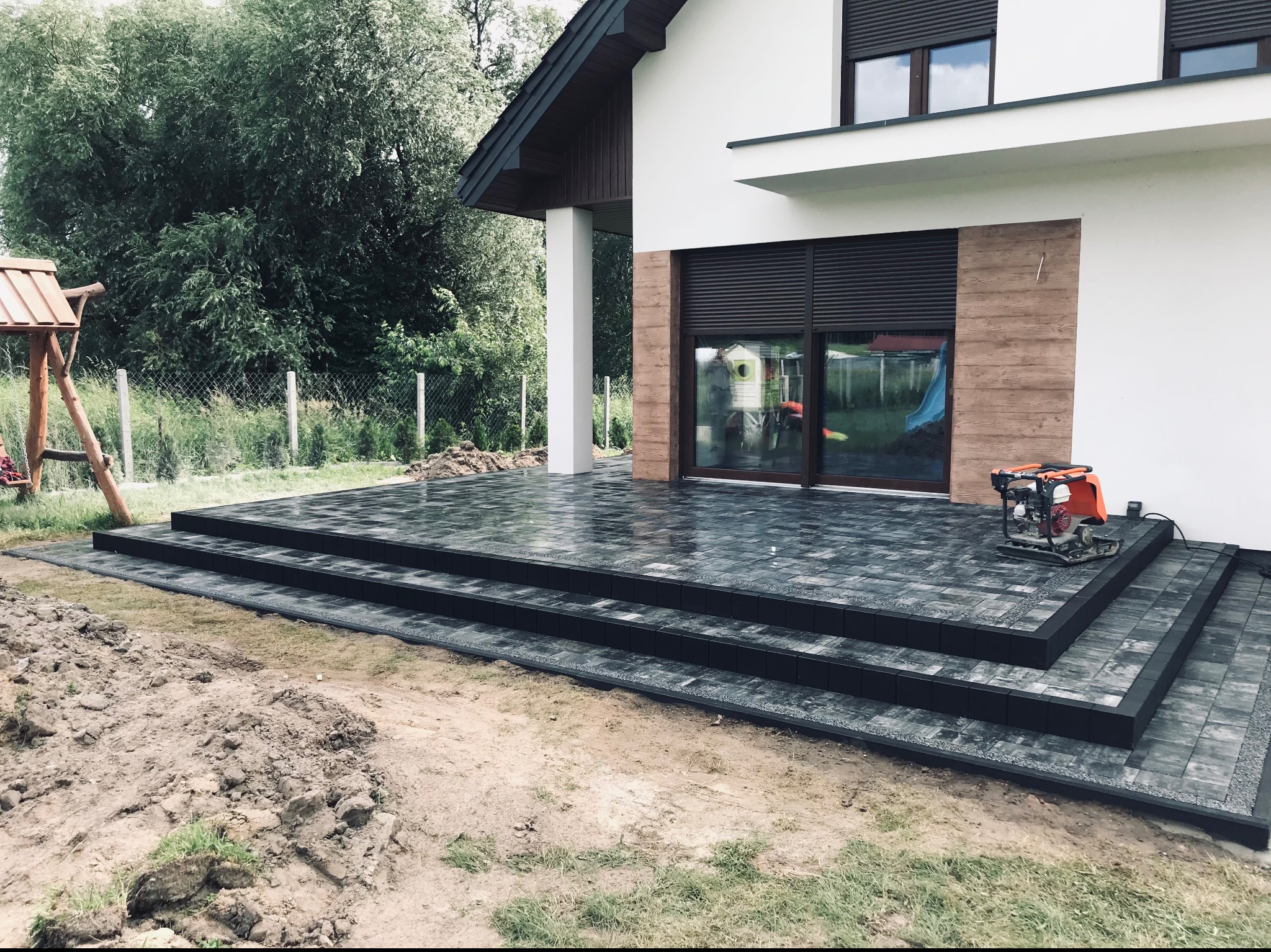 Nowoczesny taras z ciemnoszarej kostki brukowej z trzema stopniami, częściowo ukończony, z zagęszczarką w tle, Warszawa.