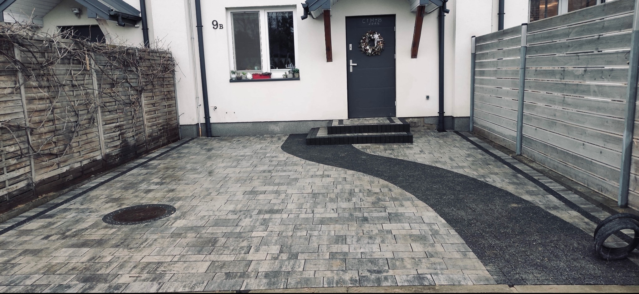 Ułożona kostka brukowa w odcieniach szarości tworząca ścieżkę prowadzącą do domu z ciemnymi schodami i dekoracyjnym wieńcem na drzwiach, obok drewniany płot i studzienka kanalizacyjna.