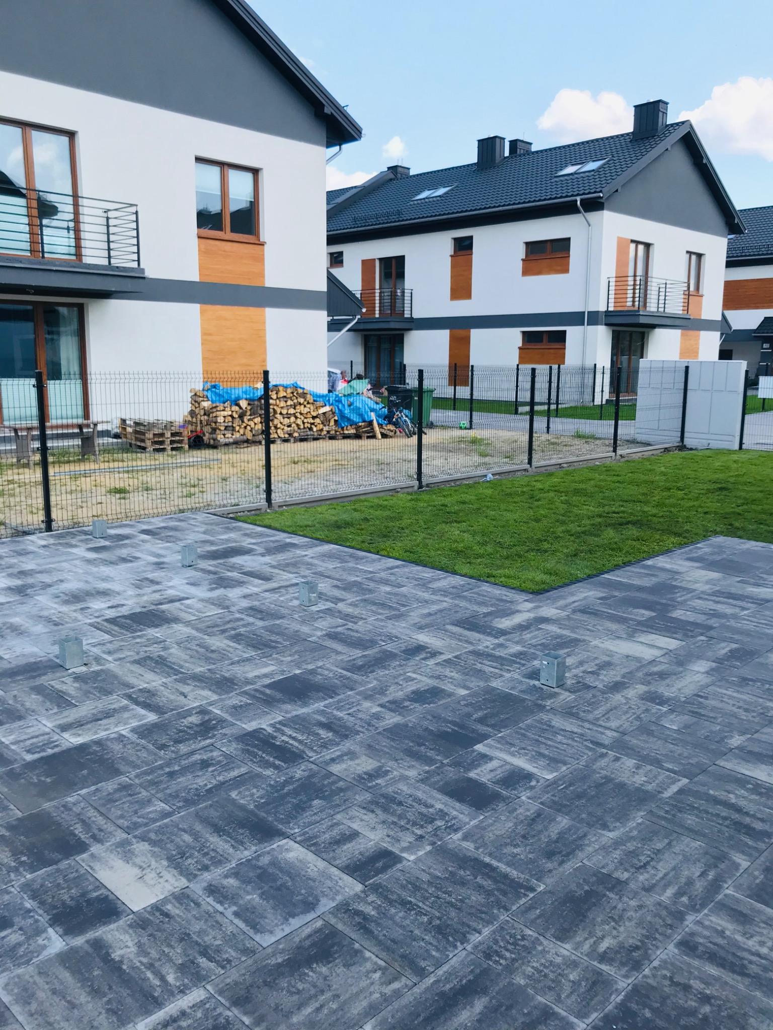 Ułożona kostka brukowa w odcieniach szarości z widocznymi metalowymi elementami montażowymi, trawnik i nowe domy w tle.