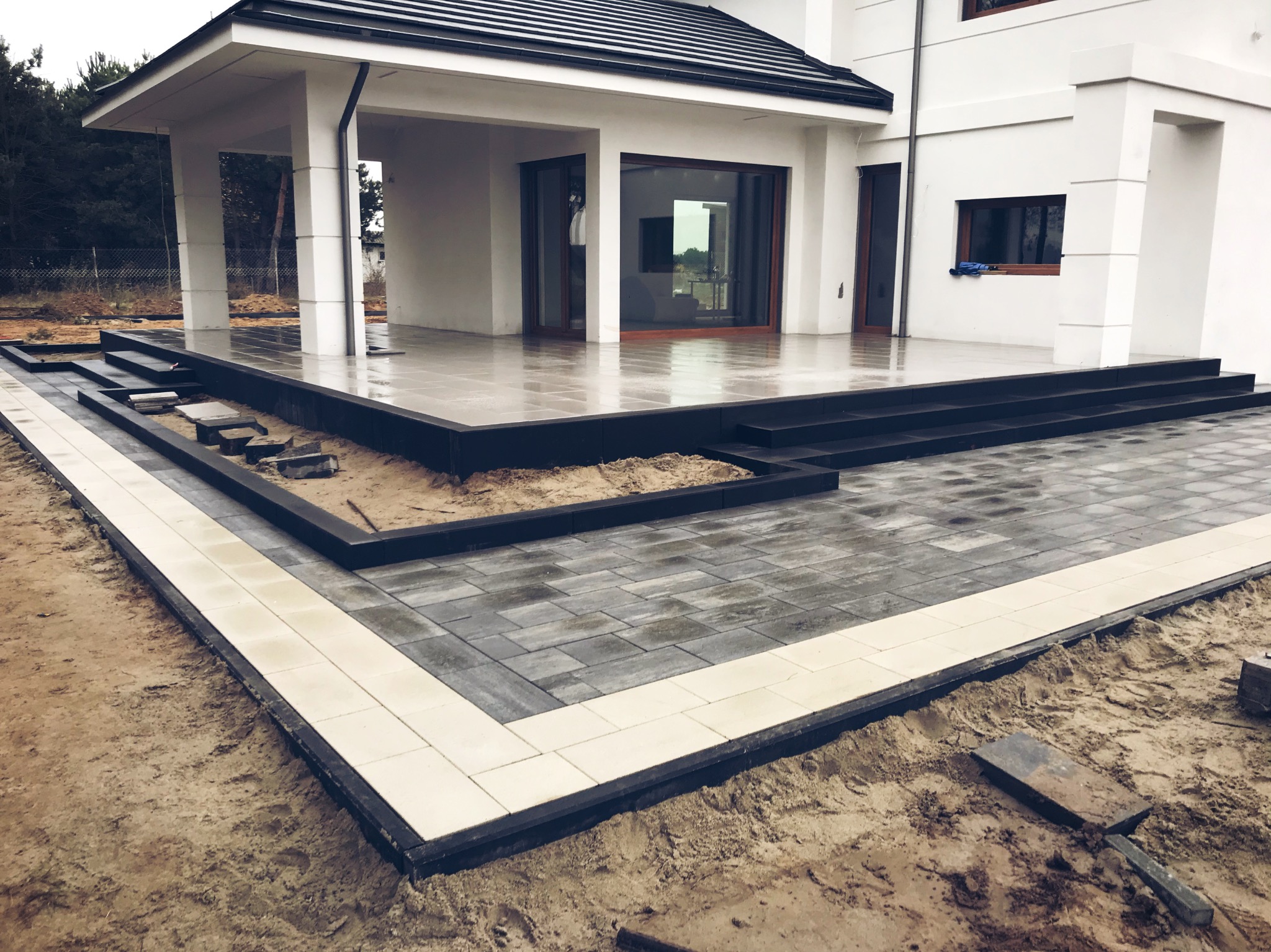 Nowoczesny taras z szarej kostki brukowej obramowany jasnymi płytami, przylegający do białego domu z dużymi oknami tarasowymi, widoczne niedokończone prace ziemne.