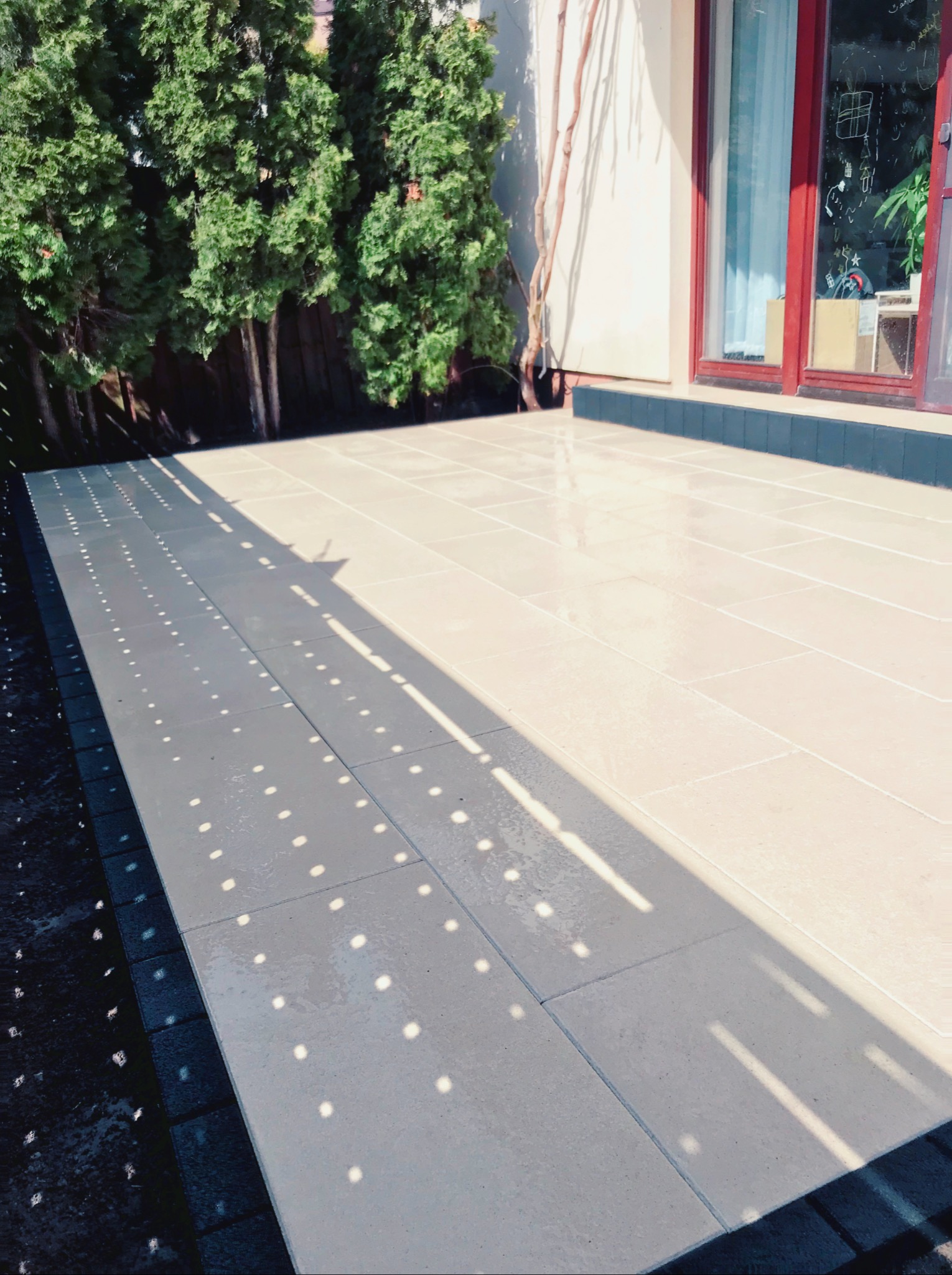 Jasna, prostokątna kostka brukowa na tarasie, oświetlona promieniami słońca tworzącymi wzór kropek, z ciemnym obramowaniem i zielonymi krzewami w tle.