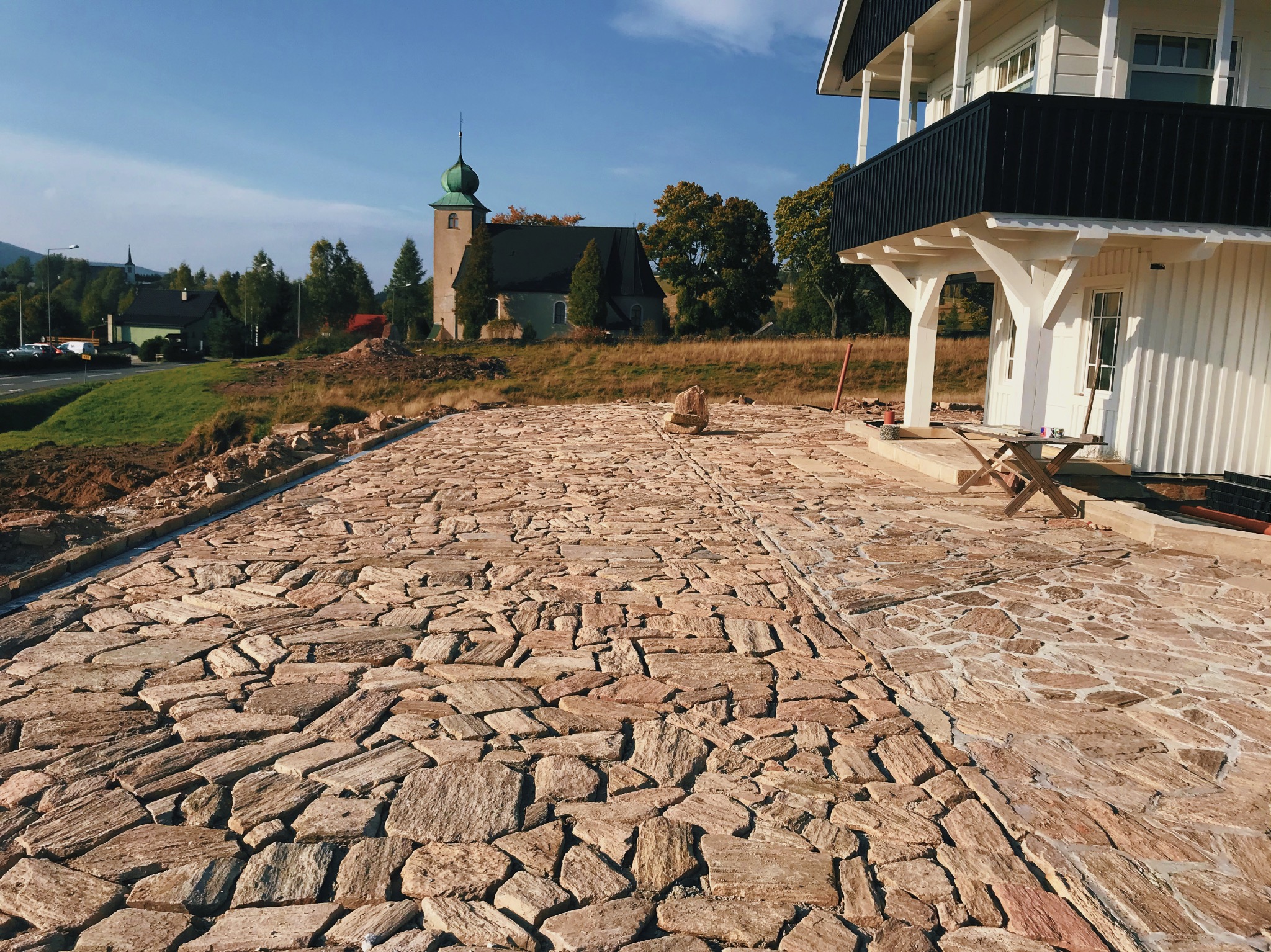 Nierówna powierzchnia z nieregularnych, brązowo-beżowych kamieni tworząca ścieżkę lub podjazd w naturalnym otoczeniu z widokiem na kościół i dom z balkonem.