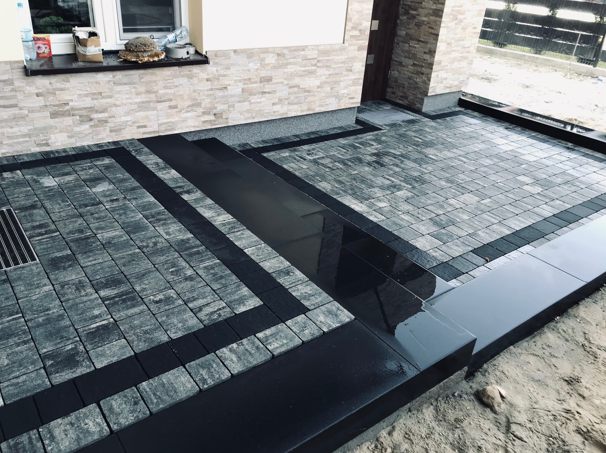 Ułożona kostka brukowa w odcieniach szarości z czarnymi obramowaniami, widoczny odpływ liniowy i fragment elewacji budynku z oknem.