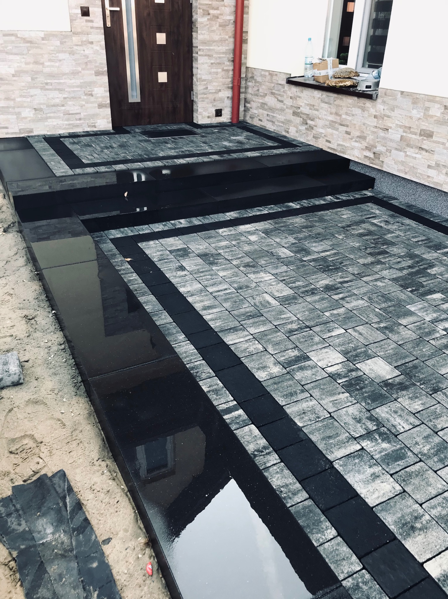 Nowoczesne wejście do domu z kostki brukowej w odcieniach szarości, z czarnymi obrzeżami i stopniami. Widoczne dwa poziomy wejścia oraz odbicie budynku w mokrej powierzchni.