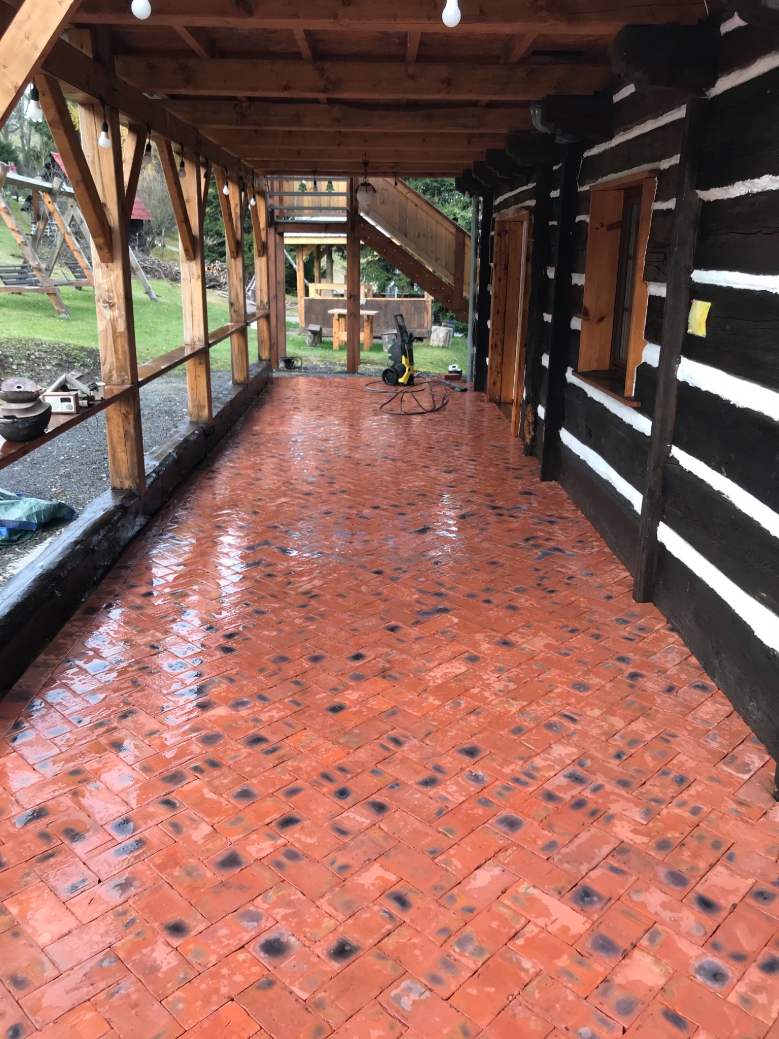 Czerwona, nowo ułożona kostka w jodełkę na zadaszonym tarasie rustykalnego domu z bali, z widocznym sprzętem do czyszczenia ciśnieniowego.