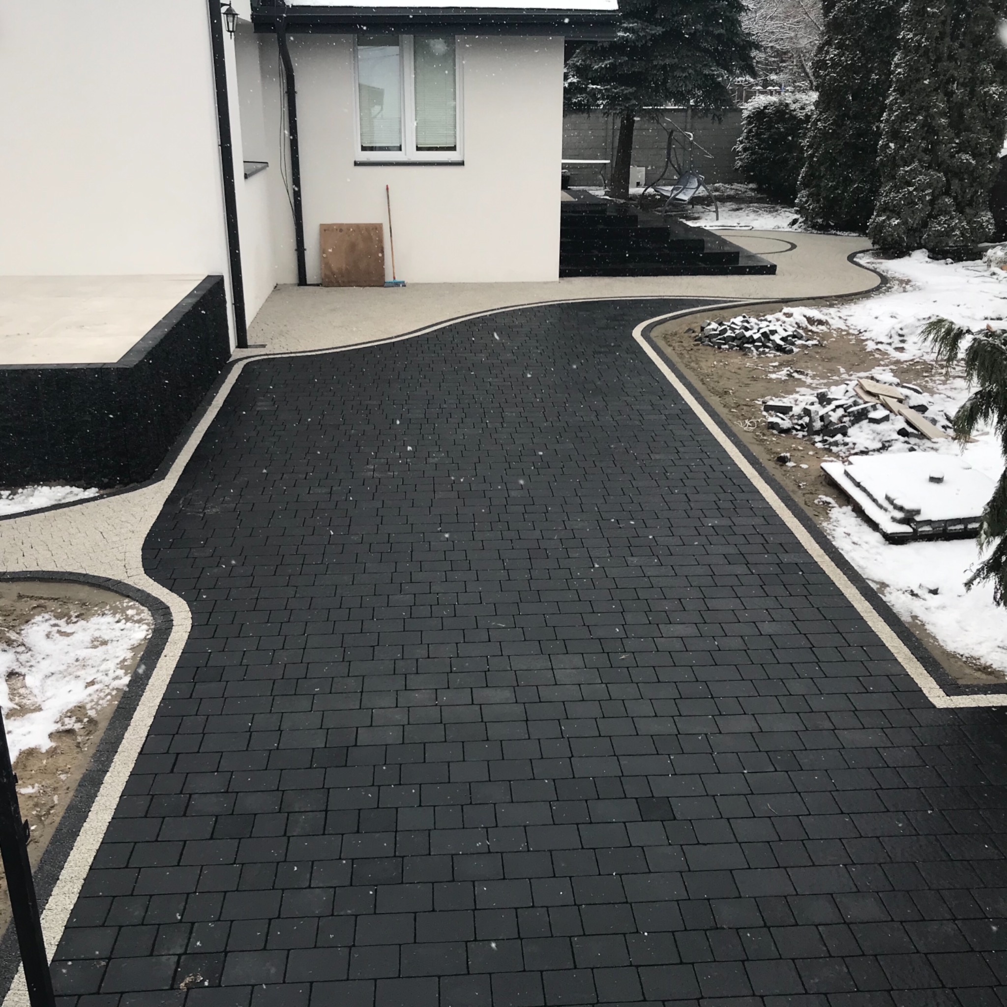 Nowoczesny podjazd z ciemnej kostki brukowej, obrzeża z jasnej kostki, częściowo pokryty śniegiem, widoczne elementy ogrodu i fragment domu.