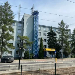 Szary budynek biurowy z niebieską konstrukcją windy zewnętrznej i antenami na dachu, widok z ulicy z torami tramwajowymi i ogrodzeniem.