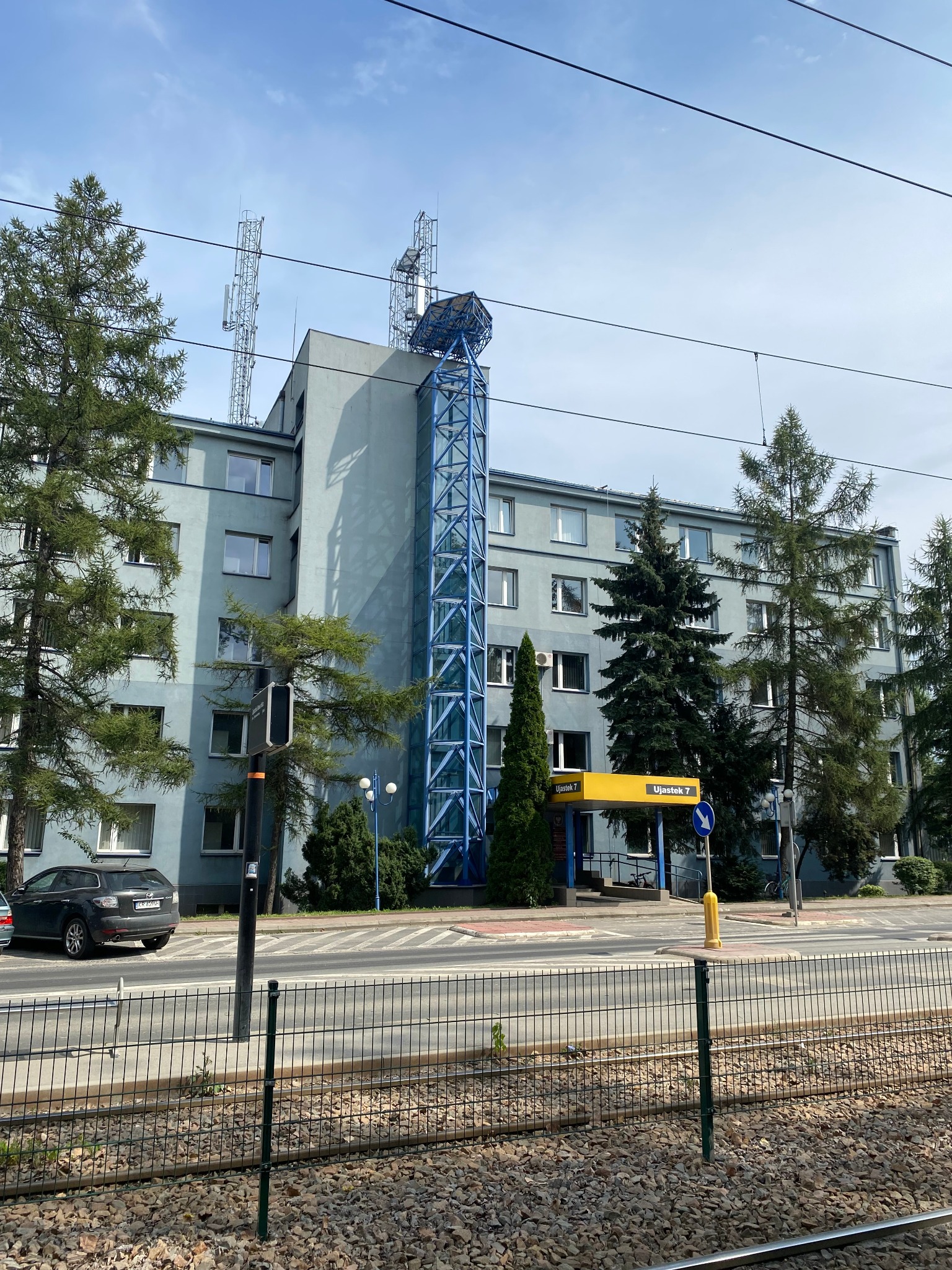 Szary budynek biurowy z niebieską konstrukcją windy zewnętrznej i antenami na dachu, widok z ulicy z torami tramwajowymi i ogrodzeniem.
