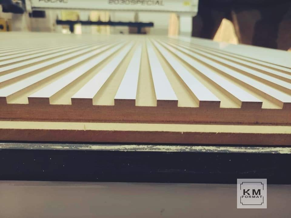 Frezowana płyta drewniana z białymi wstawkami, widoczne regularne rowki i warstwy materiału, obróbka CNC w tle.