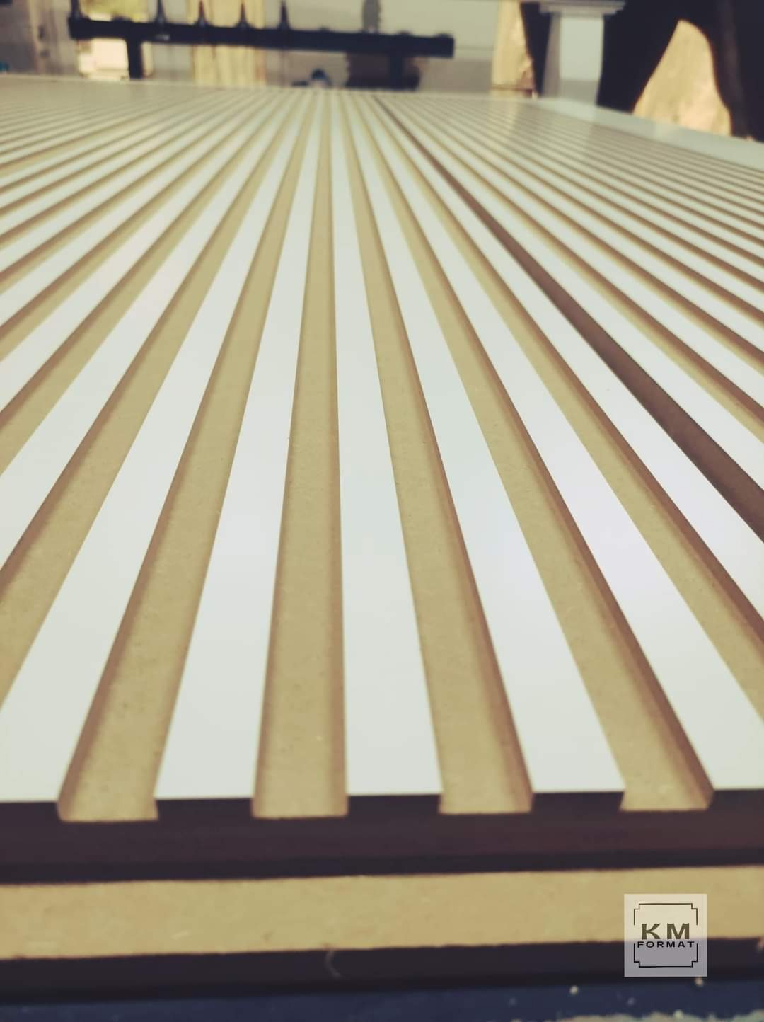 Frezowana płyta MDF z białymi wstawkami, tworząca regularny wzór rowków, widoczna z niskiej perspektywy, z logo firmy w prawym dolnym rogu.