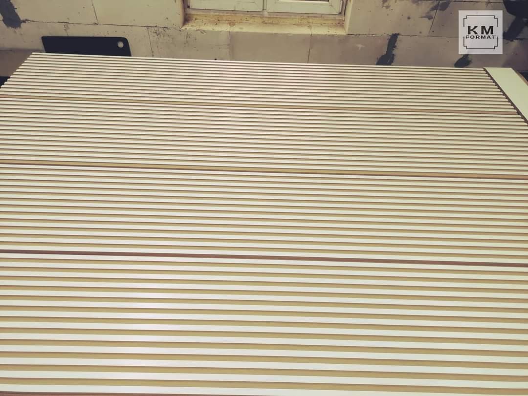 Białe i drewniane lamele ułożone równolegle, widoczne na tle surowych murów i okna, potencjalnie element szafy lub dekoracji ściennej w trakcie montażu.