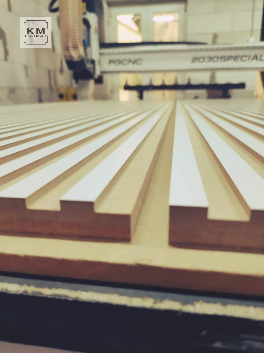 Frezowanie płyty drewnianej z wyfrezowanymi podłużnymi rowkami na maszynie CNC, widoczne logo firmy KM FORMAT w lewym górnym rogu.