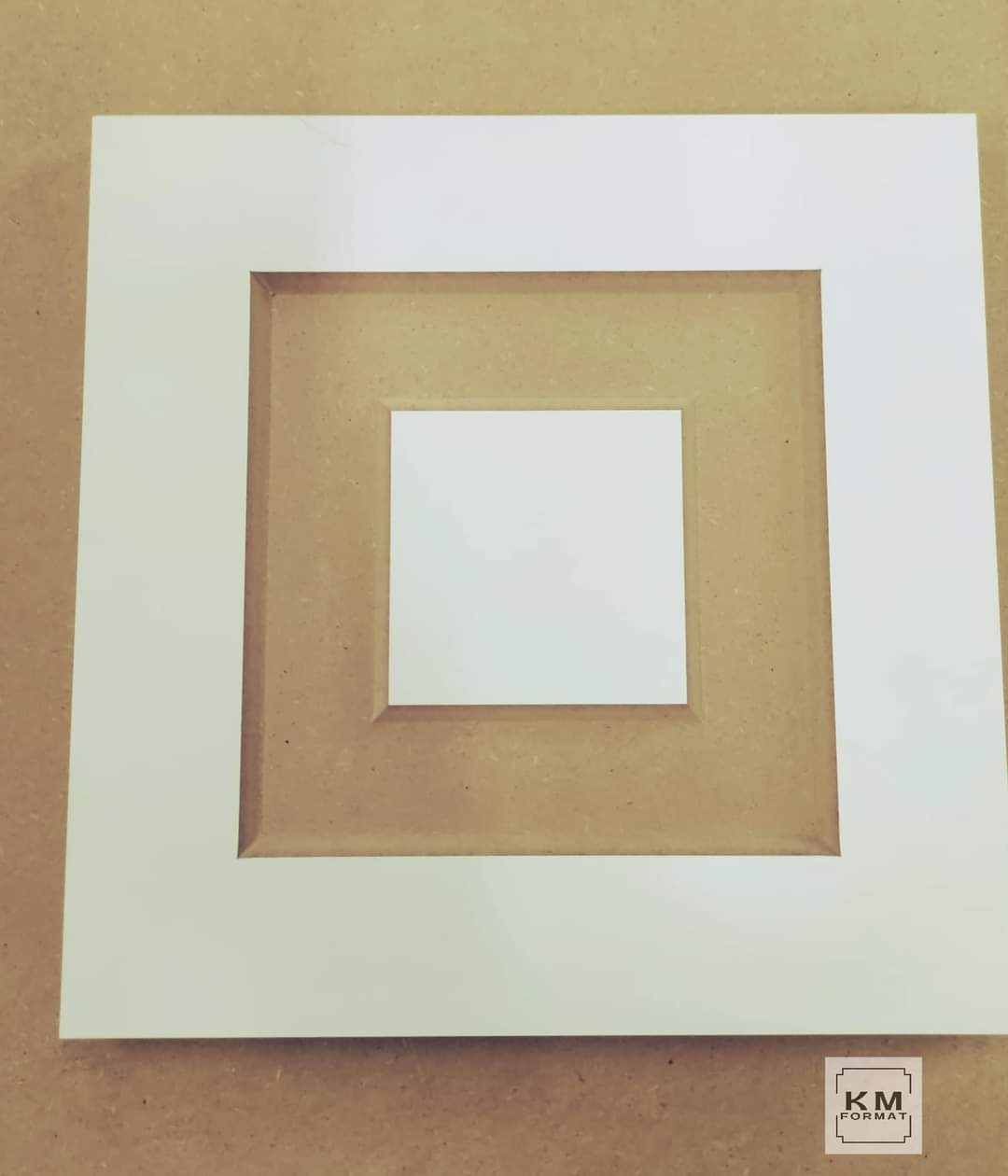 Biała, kwadratowa ramka z frezowanym profilem, na brązowym tle płyty stolarskiej, widok z góry.