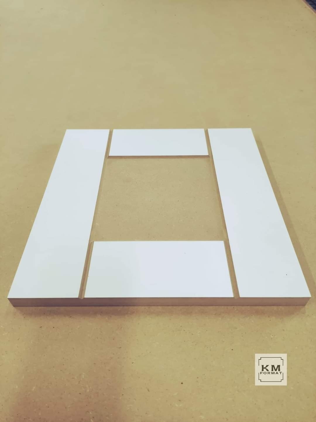 Elementy ramy z białego drewna, prawdopodobnie do drzwi lub frontu meblowego, ułożone na stole warsztatowym przed sklejeniem.