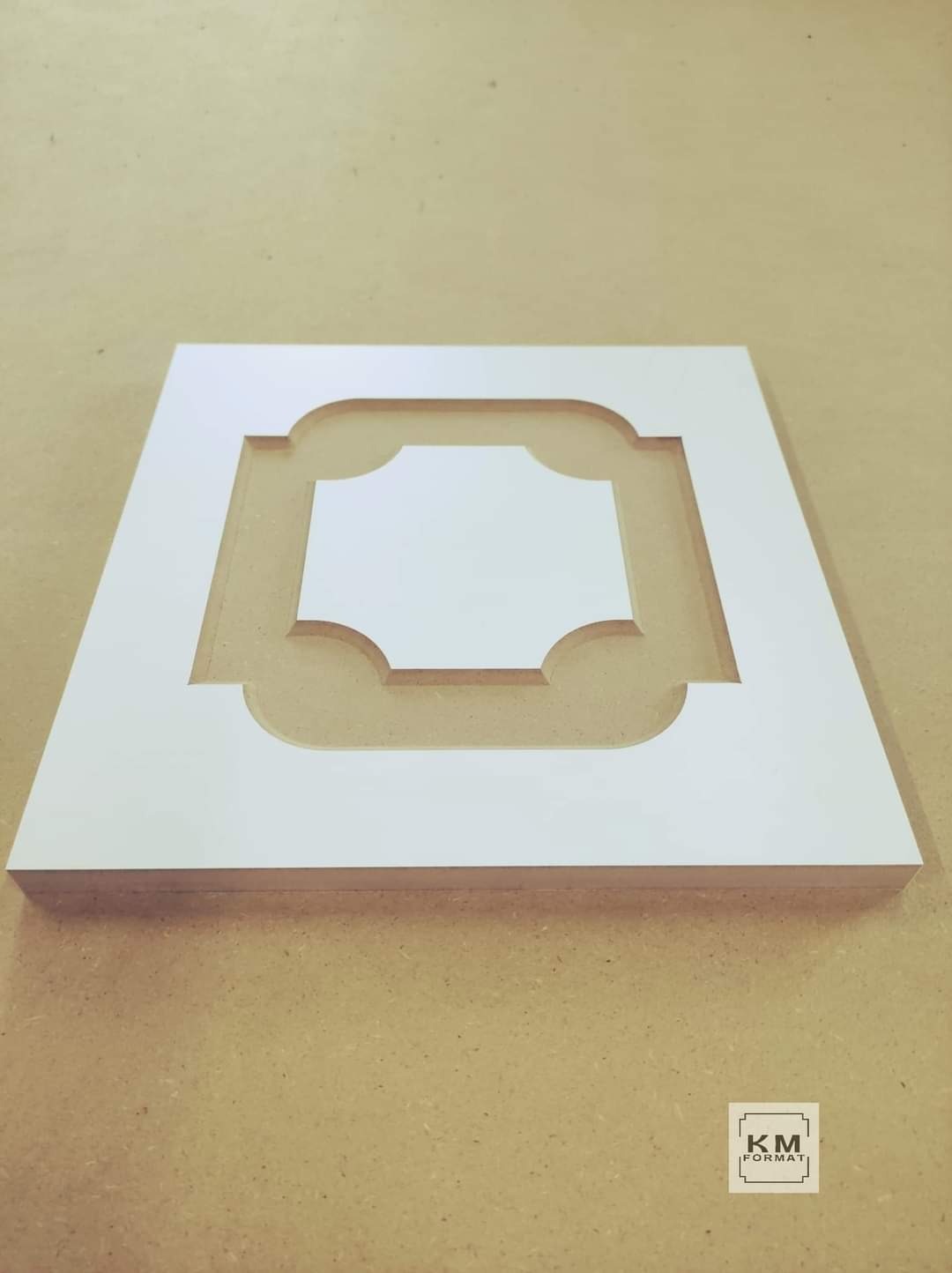 Biały, kwadratowy panel dekoracyjny z wyciętym geometrycznym wzorem, leżący na powierzchni przypominającej płytę stolarską. Widoczne logo firmy 'KM FORMAT' w prawym dolnym rogu.