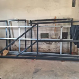 Elementy konstrukcyjne bramy przesuwnej i ogrodzenia panelowego w warsztacie, widoczne spawane ramy metalowe i fragmenty siatki ogrodzeniowej.