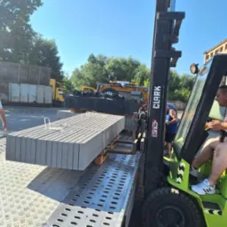 Operator w wózku widłowym marki Clark transportuje paletę z prefabrykowanymi elementami betonowymi, prawdopodobnie do budowy ogrodzenia, na platformie transportowej; w tle stoi pracownik i ciężarówka.