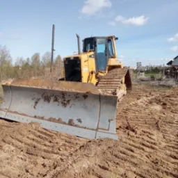 Żółta spycharka Caterpillar BOSS 9+20 z brudnym lemieszem stoi na nierównym, gliniastym terenie, w tle dom i drzewa pod błękitnym niebem.