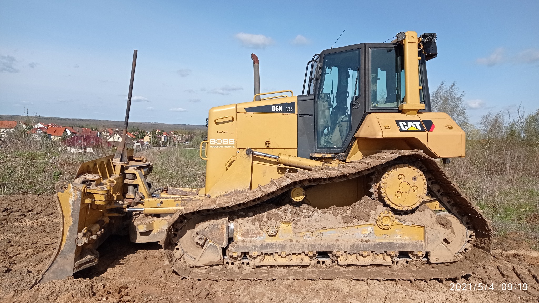 Żółty spychacz marki CAT D6N LGP, z widocznymi śladami błota na gąsienicach i lemieszu, stoi na terenie budowy z zabudowaniami w tle, pod błękitnym niebem z lekkimi chmurami.