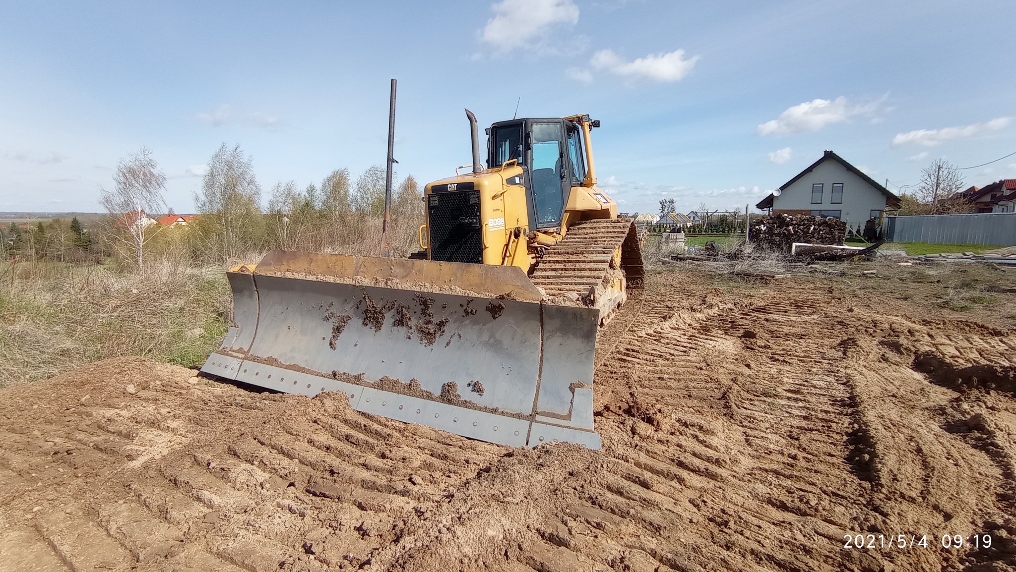 Żółta spycharka Caterpillar BOSS 9+20 z brudnym lemieszem stoi na nierównym, gliniastym terenie, w tle dom i drzewa pod błękitnym niebem.