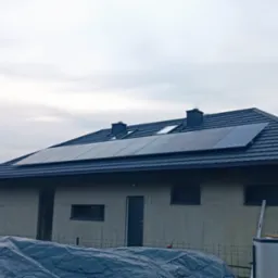 Niedokończony dom jednorodzinny z zamontowanymi panelami słonecznymi na dachu pokrytym ciemną dachówką, widoczne dwa kominy i okna dachowe, ściany domu w stanie surowym, przykryte folią elementy...