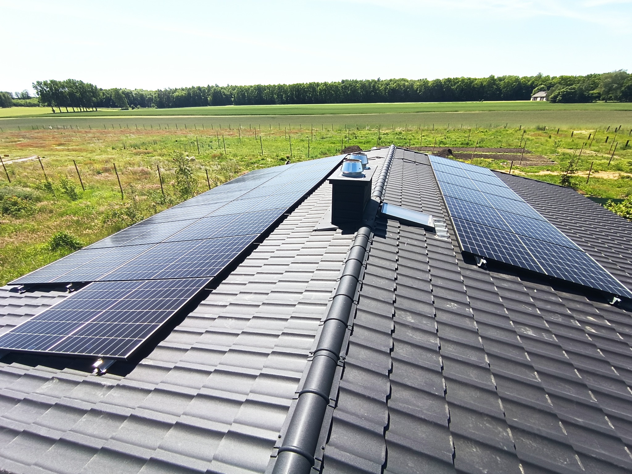 Panele fotowoltaiczne zamontowane na ciemnoszarym dachu z dachówki ceramicznej, widok z góry na tle zielonego pola i lasu.