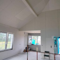 Usługi ogólnobudowlane Artur Zieliński - Wnętrze w trakcie wykańczania: ściany i skośny sufit pokryte płytami gipsowo-kartonowymi, widoczne okna z widokiem na zewnątrz, w tle błękitna toaleta przenośna.