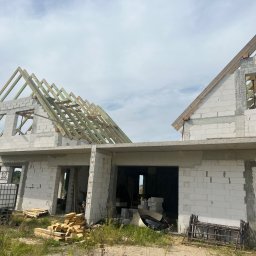 Usługi ogólnobudowlane Artur Zieliński - Surowy stan budowy bliźniaka z widoczną drewnianą konstrukcją dachu w trakcie montażu, ściany z białego bloczka, teren wokół zarośnięty trawą, w tle błękitne niebo z chmurami.