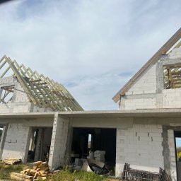 Usługi ogólnobudowlane Artur Zieliński - Budowa domu jednorodzinnego w stanie surowym otwartym z widoczną konstrukcją dachu i ścianami z bloczków betonowych oraz stosem drewna i rusztowaniem na ziemi.