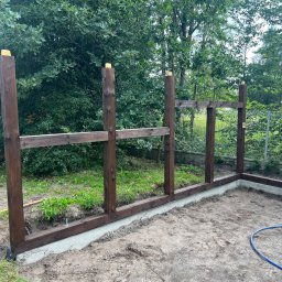 Usługi ogólnobudowlane Artur Zieliński - Wczesny etap budowy konstrukcji z ciemnego drewna, prawdopodobnie altany lub wiaty, na betonowej podstawie. W tle widoczne drzewa i krzewy.