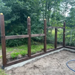 Wczesny etap budowy konstrukcji z ciemnego drewna, prawdopodobnie altany lub wiaty, na betonowej podstawie. W tle widoczne drzewa i krzewy.