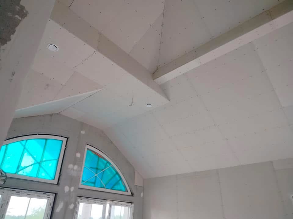 Sufit poddasza wykończony płytami gipsowo-kartonowymi, widoczne belki konstrukcyjne i okrągłe lampy sufitowe, dwa okna o nietypowym kształcie z niebieskimi szybami.