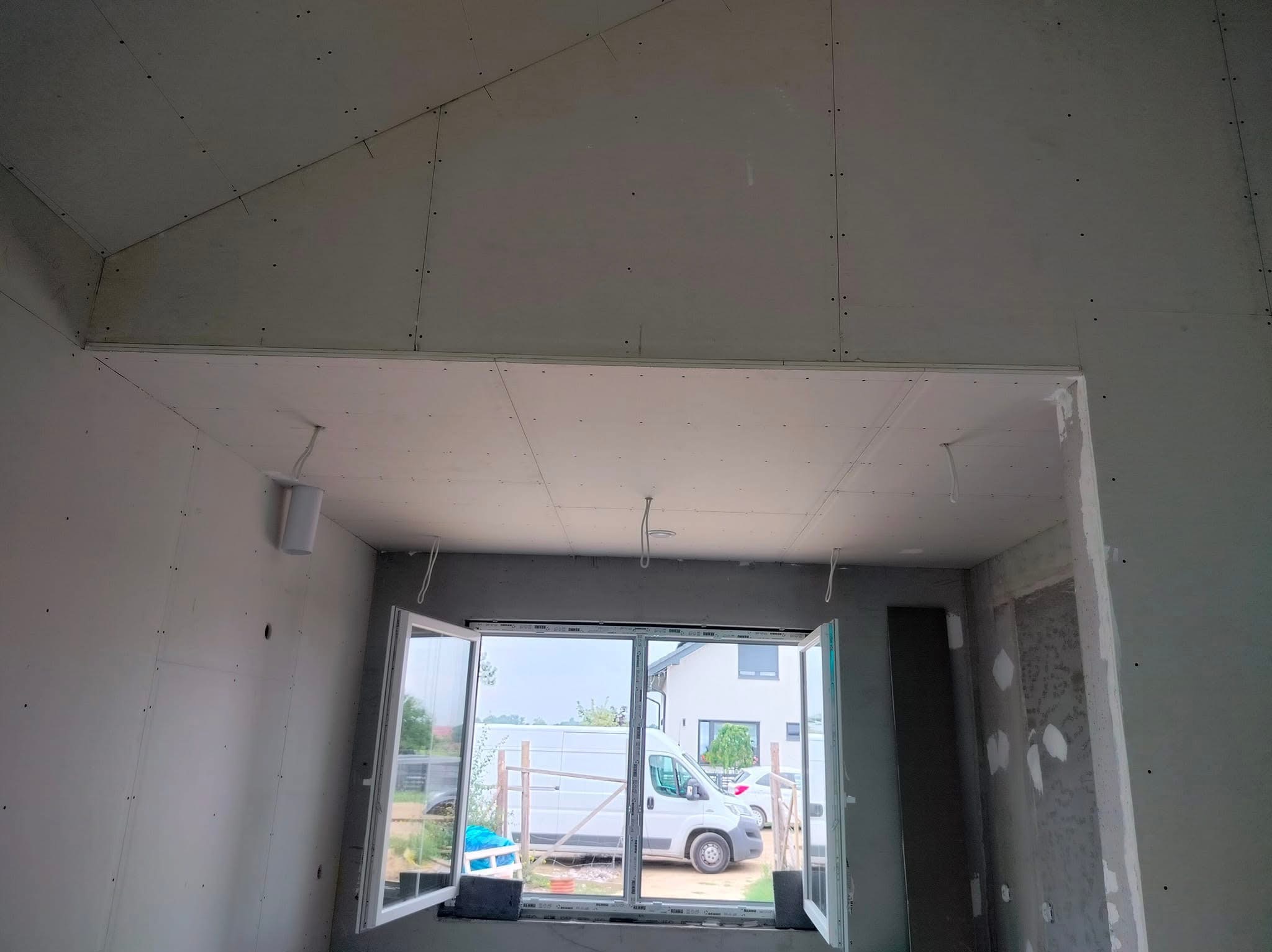 Wnętrze pokoju w trakcie wykańczania z zamontowanymi płytami gipsowo-kartonowymi na ścianach i suficie, widok przez otwarte okno na podwórko z zaparkowanym białym vanem.