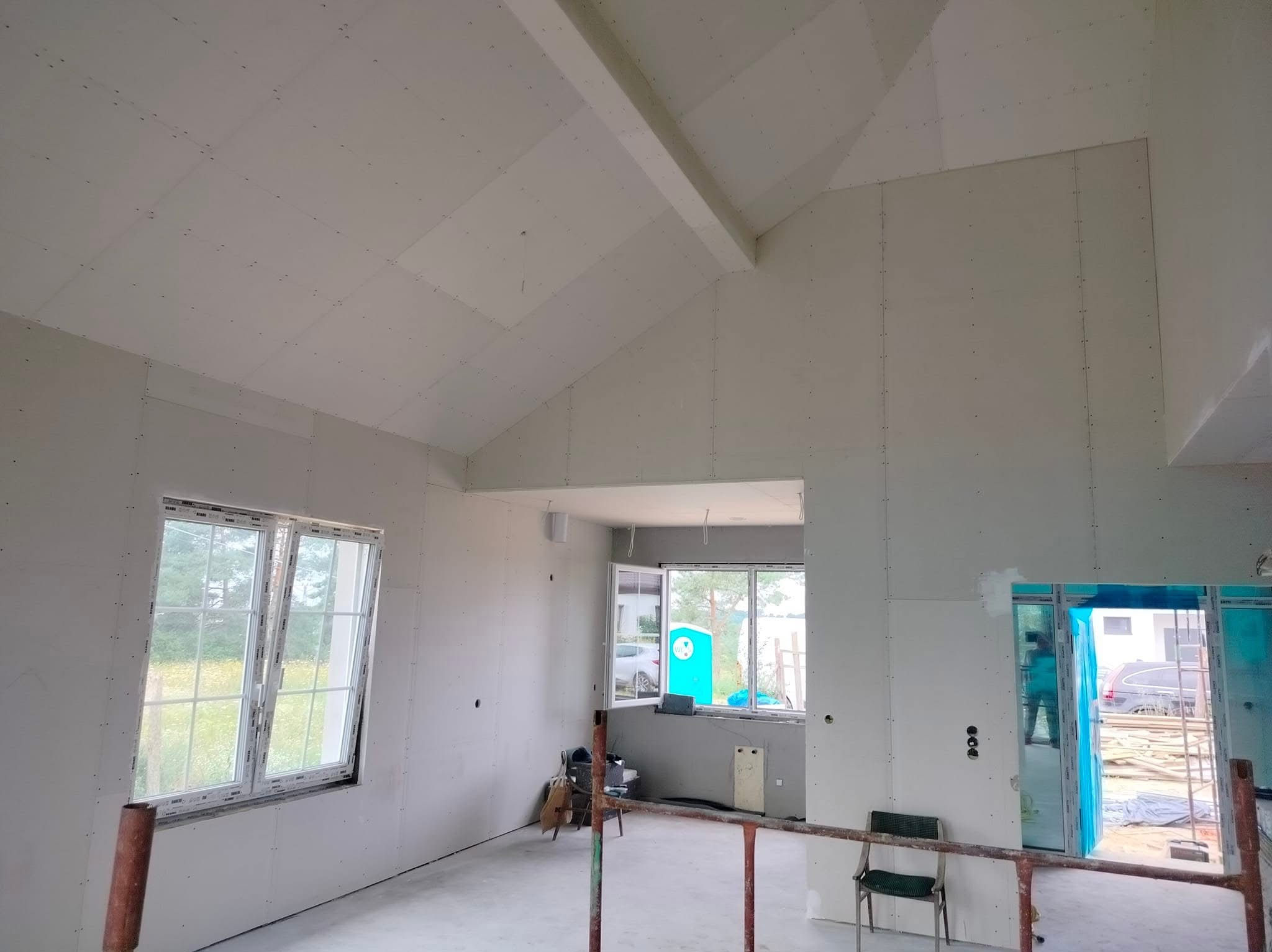 Wnętrze w trakcie wykańczania: ściany i skośny sufit pokryte płytami gipsowo-kartonowymi, widoczne okna z widokiem na zewnątrz, w tle błękitna toaleta przenośna.