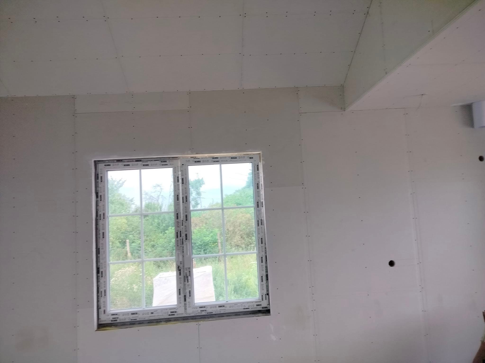 Wnętrze pokoju w trakcie wykańczania, ściany i sufit pokryte płytami gipsowo-kartonowymi, widoczne ślady po wkrętach, okno PCV z widokiem na zieleń na zewnątrz.