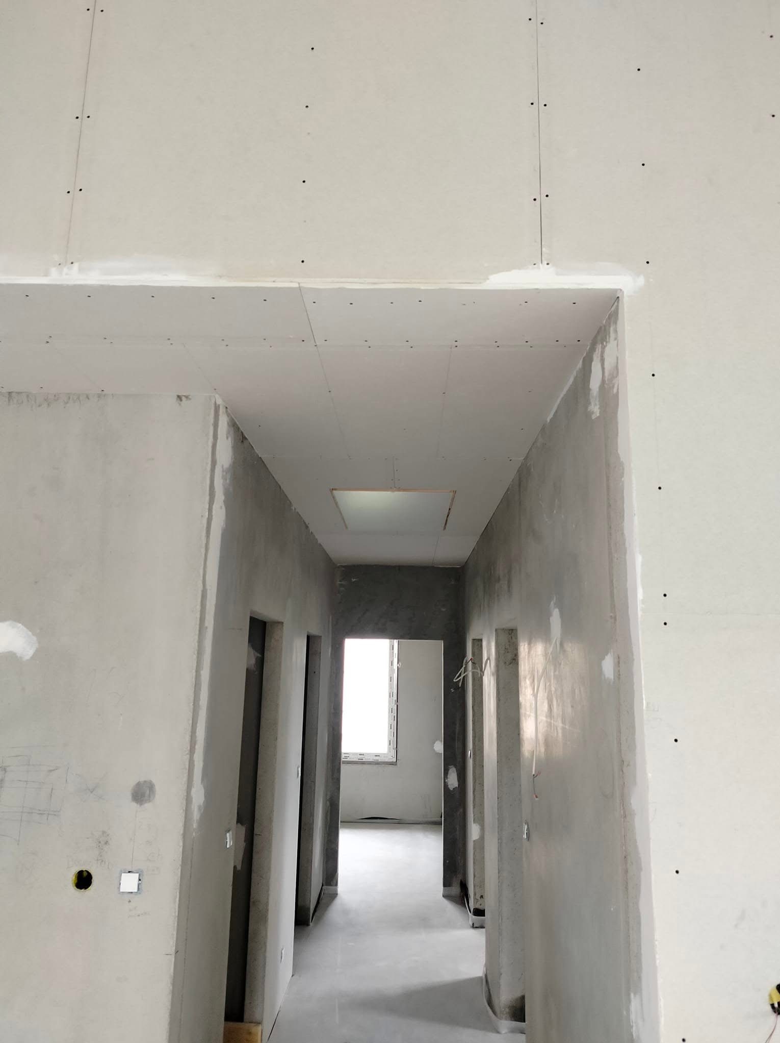 Wnętrze w trakcie wykańczania: surowe ściany z płyt gipsowo-kartonowych, widoczne łączenia i ślady szpachlowania, przygotowane otwory na instalacje elektryczne, sufit z otworem rewizyjnym.