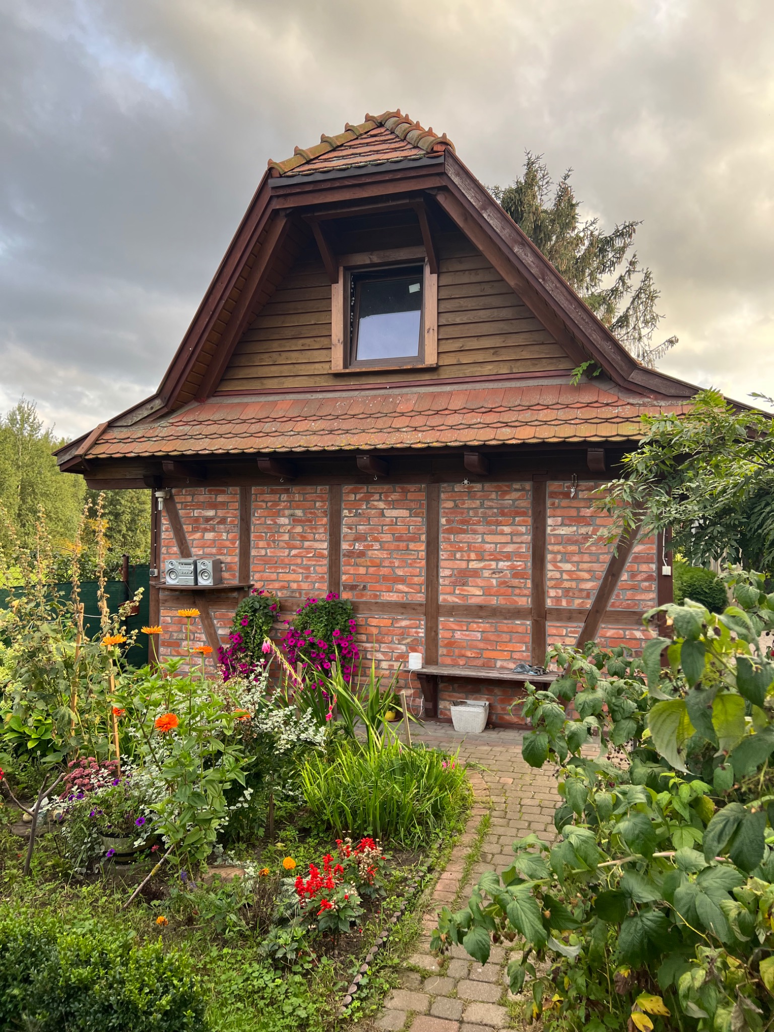 Niewielki budynek z czerwonej cegły i drewnianą konstrukcją szkieletową, z dachem pokrytym czerwoną dachówką, otoczony bujnym ogrodem kwiatowym i ścieżką z kostki.