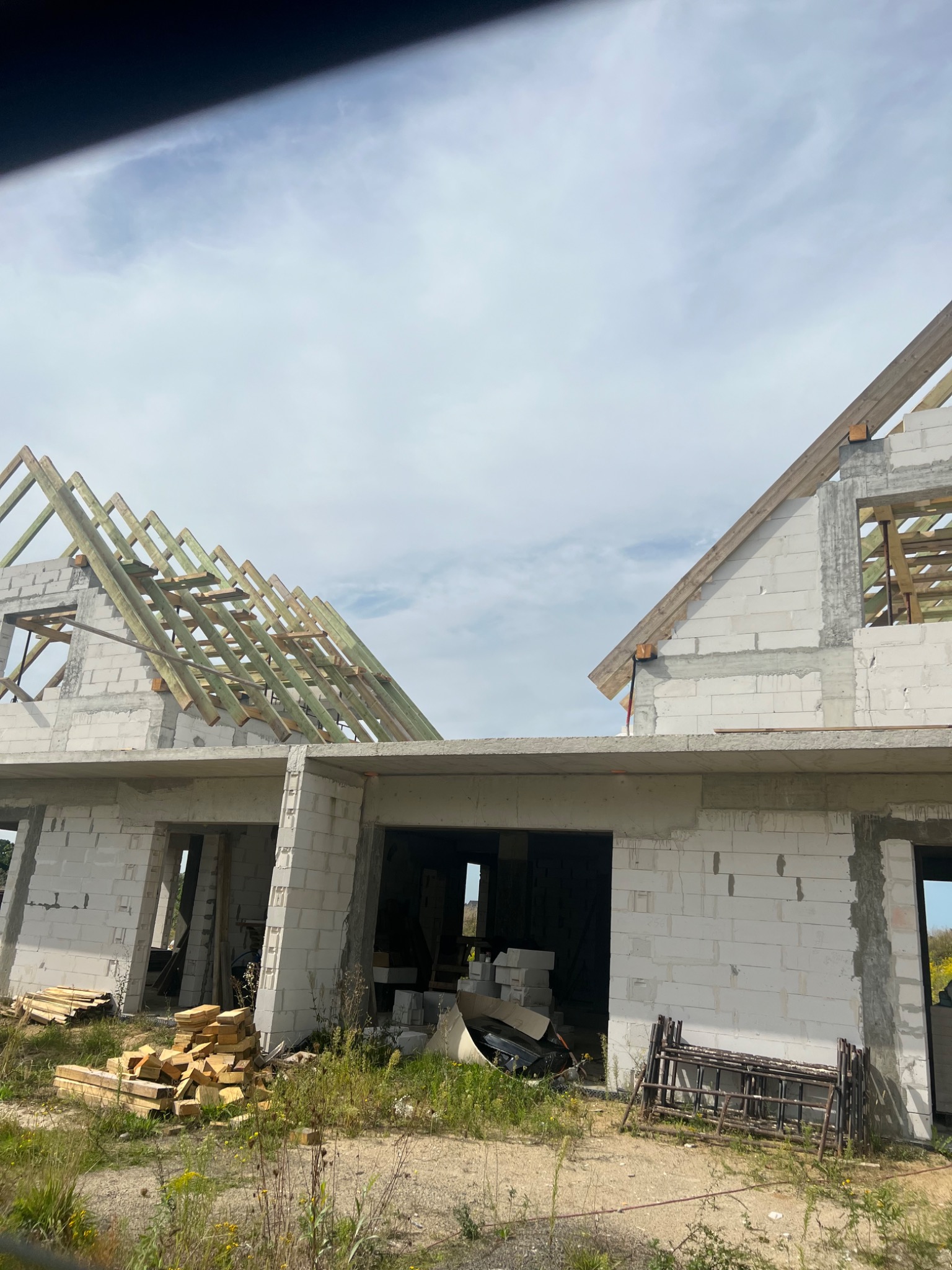 Budowa domu jednorodzinnego w stanie surowym otwartym z widoczną konstrukcją dachu i ścianami z bloczków betonowych oraz stosem drewna i rusztowaniem na ziemi.